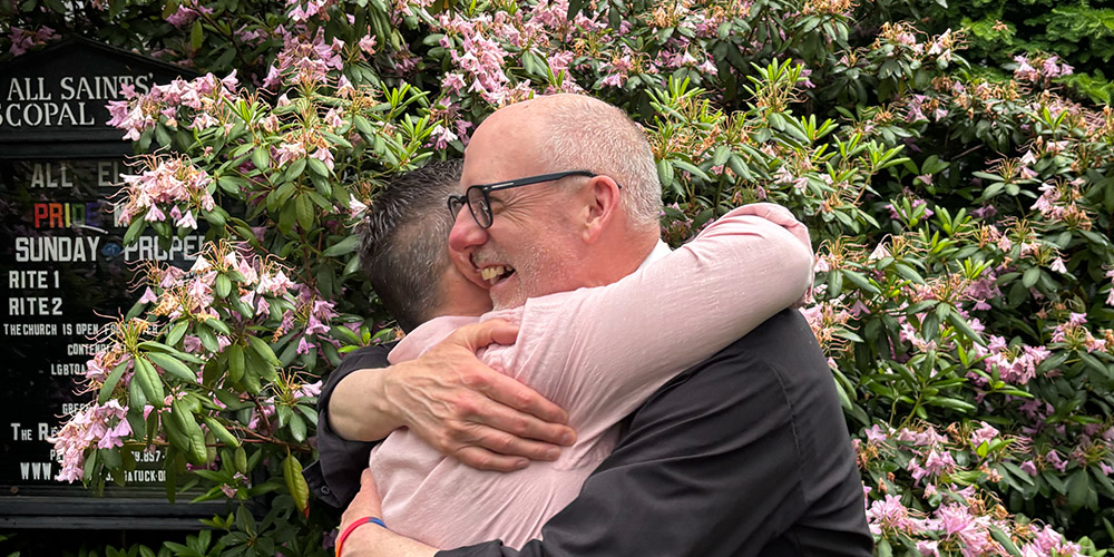 Two people hug and smile outside All Saints’ Episcopal Church, surrounded by pink flowering bushes.
