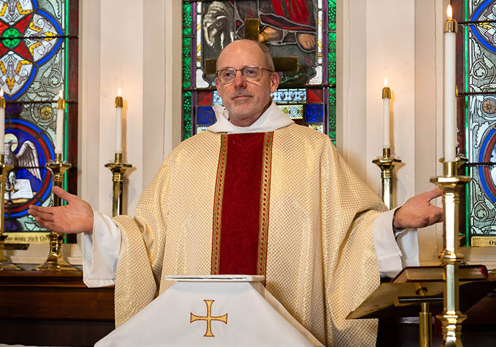 700px_media-block-set-III_0000__The Episcopal Church VIII A priest in cream-colored vestments stands at the altar with arms open, surrounded by lit candles and stained-glass windows.