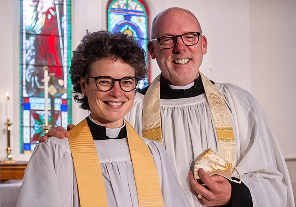 Two smiling clergy members in white robes and gold stoles stand together in front of stained-glass windows