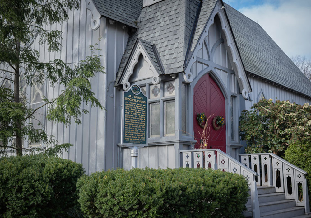 Exterior of All Saints’ Episcopal Church with red front doors, steps, shrubs, and a church sign near the entrance.