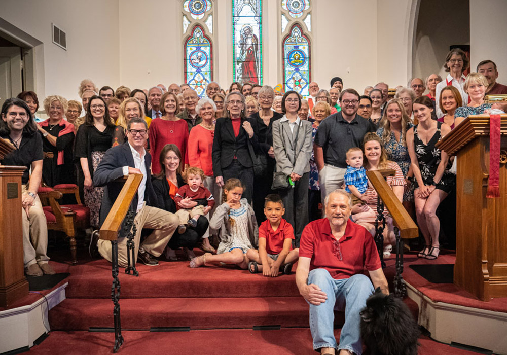 700px_media-block_0001__community life A large multigenerational church group poses together on the steps inside a sanctuary beneath stained-glass windows.