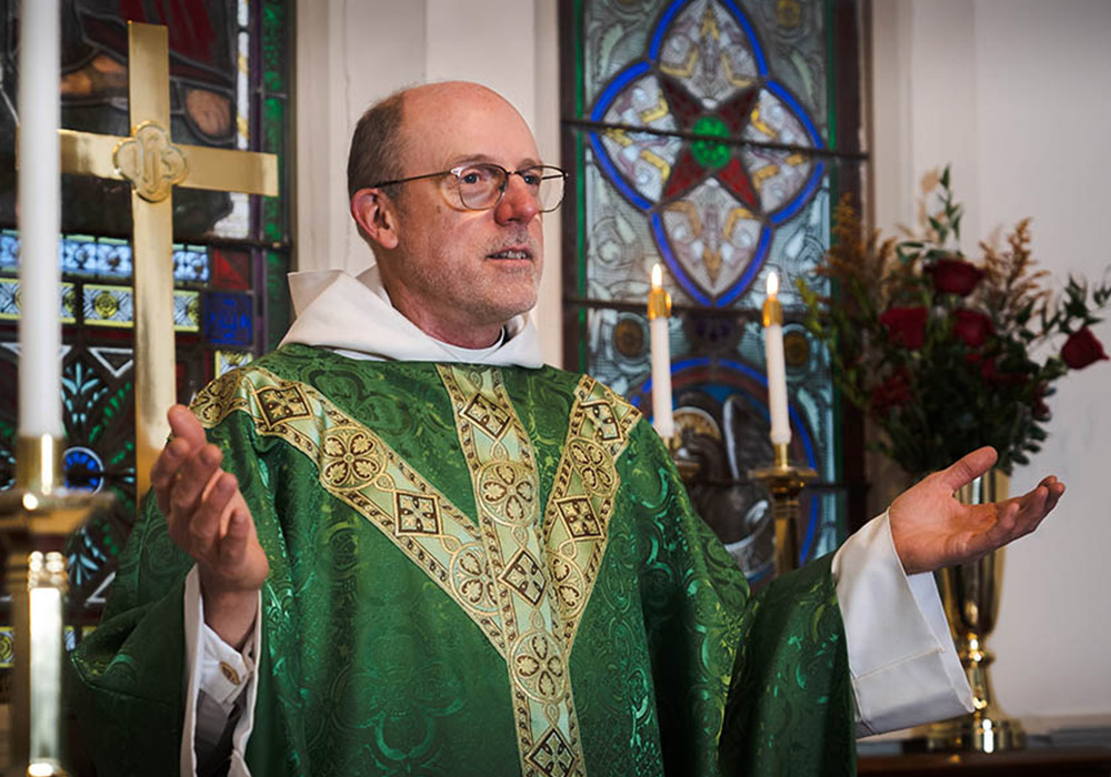 A priest in green vestments stands near candles and stained-glass windows with hands open in prayer.