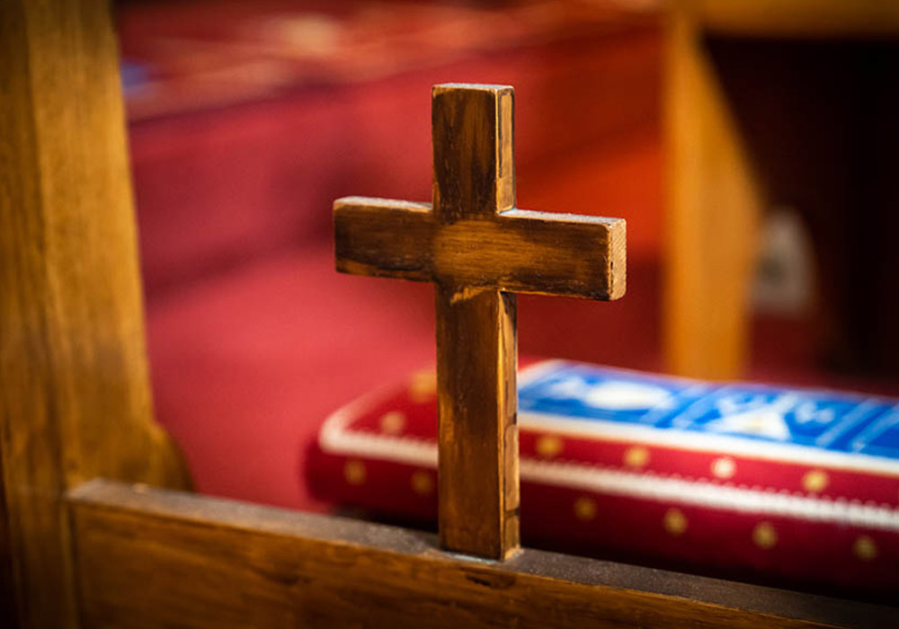 700px_media-block_0008__The Episcopal Church II Close-up of a small wooden cross on a church pew, with a red kneeler blurred in the background.