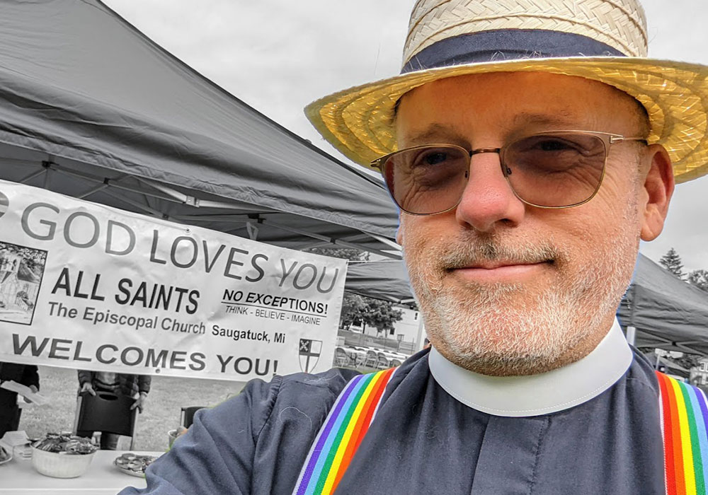 smiling priest in sunglasses and a straw hat poses for a selfie beside an All Saints Episcopal Church sign that reads “God loves you” and “Welcomes you.”